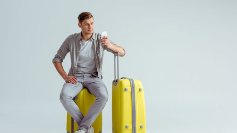 Ein junger Mann auf Reisen Ein jungern Mann sitzt auf einem gelben Koffer und schaut auf sein Smartphone in seiner linken Hand. Seinen linken Oberarm hat er auf den Griff eines weiteren gelben Koffer gestützt.