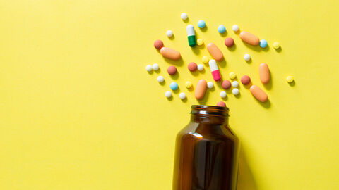 Nahrungsergänzungsmittel Eine braune kleine Glasflasche liegt auf der Seite auf einem gelben Untergrund. Mehrere runde, ovale und unterschiedlich bunte Tabletten liegen vor der Flaschenöffnung verteilt.